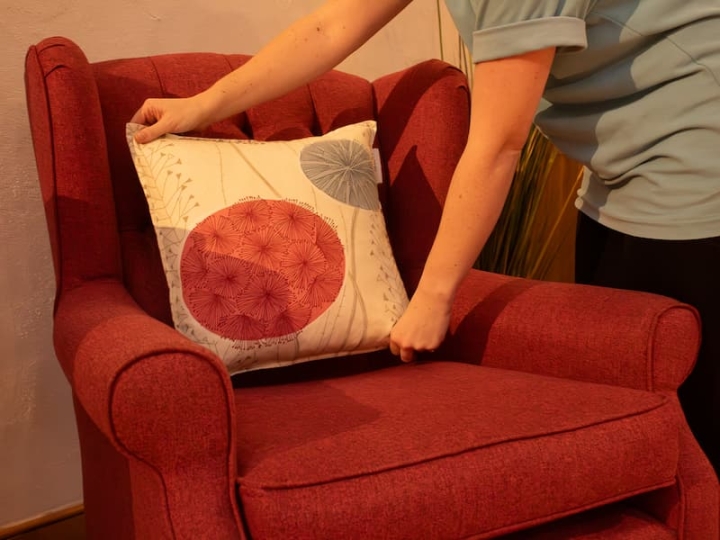 A cleaning operative meticulously placing a freshly cleaned cushion on an armchair, ensuring precision and cleanliness in the arrangement.