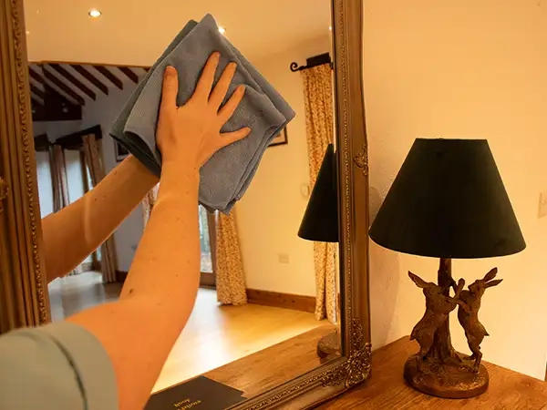 A cleaning operative meticulously cleans a mirror in a hallway within a domestic property, using a blue microfiber cloth, to ensure a hassle-free and thorough cleaning experience.