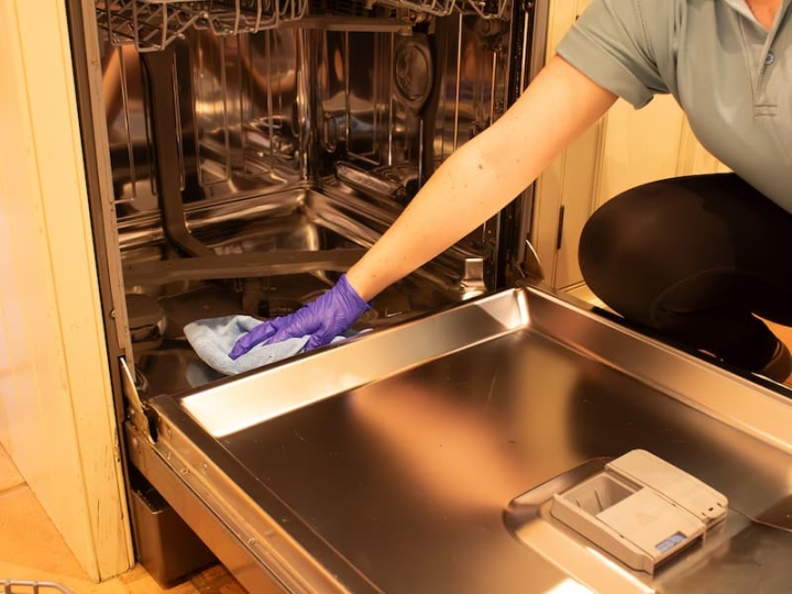 A dedicated woman, wearing a blue polo shirt with The Norfolk Cleaning Group logo, diligently cleans a dishwasher using a microfiber cloth and eco-friendly, tested products. She ensures the surface is not only clean but also disinfected, while wearing vinyl-free gloves for a meticulous and eco-conscious cleaning approach.