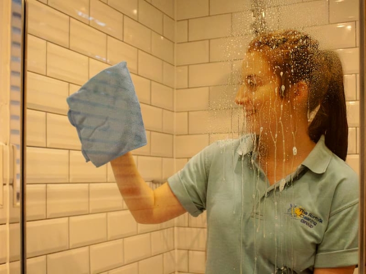 A woman diligently cleaning a shower using a blue cloth as part of the cleaning services provided by Norfolk Cleaning, ensuring a spotless and hygienic environment.
