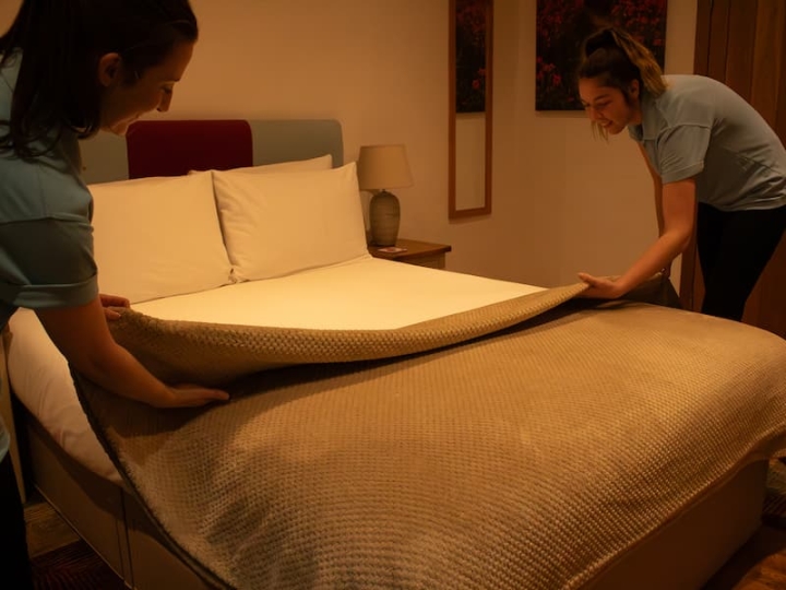 Two women skillfully making a bed with linen hire, arranging white cotton sheets meticulously. They delicately place a bed throw on top, creating a beautifully layered and inviting bed.